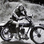 photograph of a woman in a trenchcoat and googles on a vintage bike