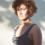 Sunlit portrait of a woman with short curly brown hair wearing a tank-top, against a white wall shadowed by a tree.