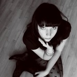 Girl in black dress on wood flooring looking up at the camera.