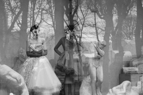 black and white photo  of two models in a window with sculptures and trees reflected in the glass