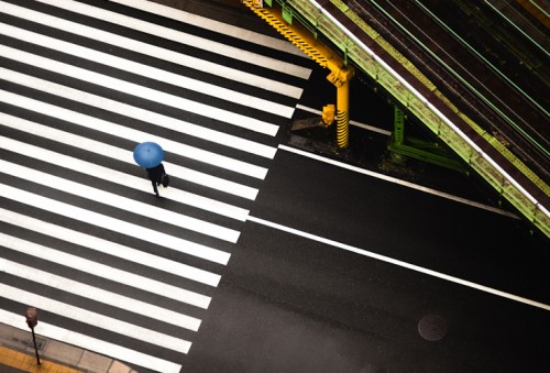 aerial photo of a man with a blue umbrella crossing zebra lines