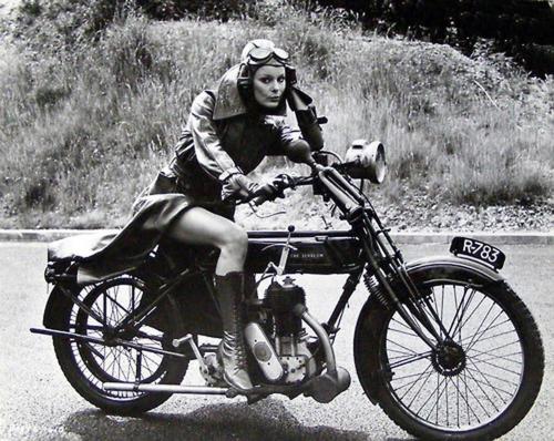 photograph of a woman in a trenchcoat and googles on a vintage bike