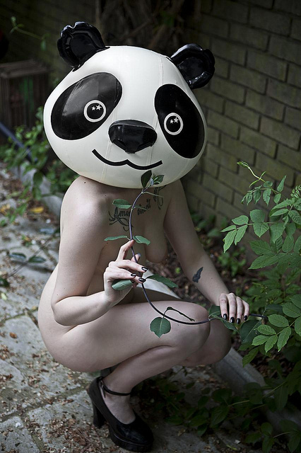 Nude girl in a blow-up panda mask crowching near a plant pretending to eat the shoots.