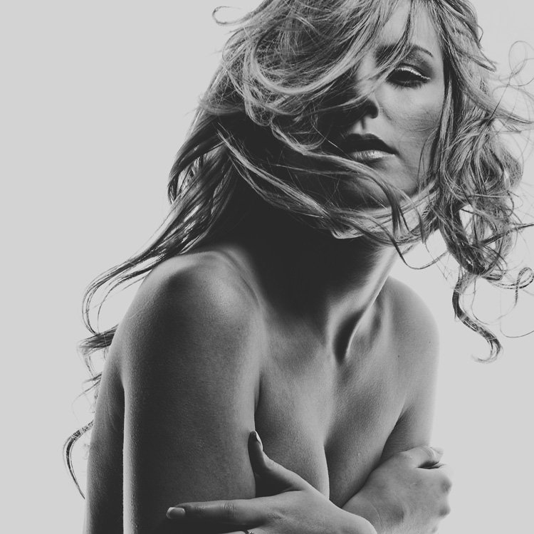 Black and white photo of topless woman with tousled blonde hair covering her breasts with her hands.