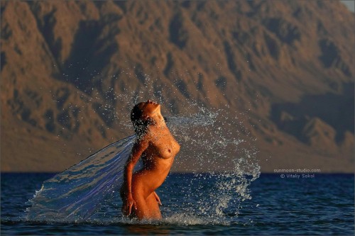 Nude woman splashing out of the water in a lagoon with rocky mountains in the background.