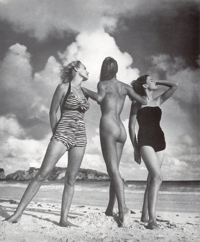 three women on a beach, two in swimsuits, one nude backing camera